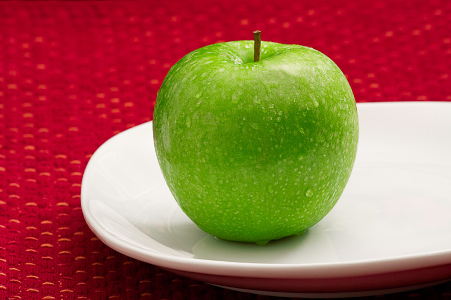 This Brian Charles Steel food photo depicts a green apple on a white plate that is sitting on a red place matt.  The apple is in the middle of the frame and takes up about a third of the photograph.  The green apple has drops of water on it giving it a fresh look.  The apple is lit from behind. The light is coming down from an elevated position in the back. 