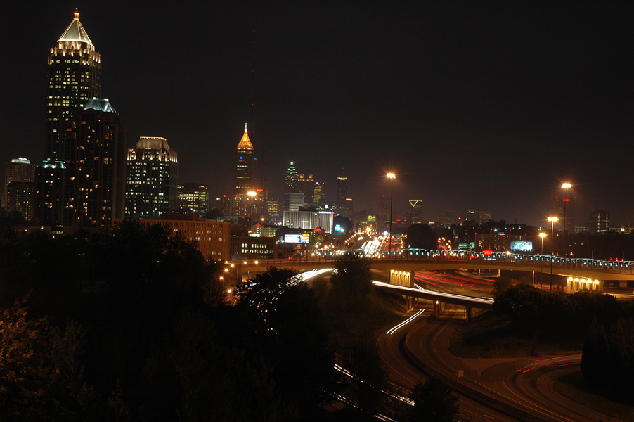 A nighttime skyline shot of midtown Atlanta. It depicts several towers along with the banana bridge and the connector.  The long exposure has caused the car lights to be beams of light. Photography by Brian Charles Steel. 