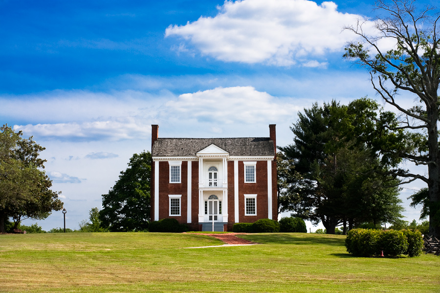 This is a Brian Charles Steel photograph of Chief Vann’s house on a sunny day in Chatsworth, Georgia.  The house is made of red brick with white pillars, and a white balcony in the middle of the second floor in the front.  The house is situated in the middle of the frame with blue sky above and short green grass below. 