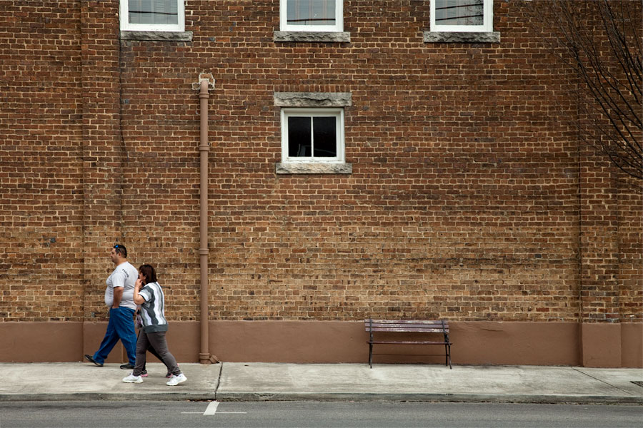 This Brian Charles Steel photograph is of the side of a brick building with white trim windows and a pipe going up the building.  The pipe’s color matches the color of the bricks.  Towards the bottom of the photograph is a sidewalk with a park bench.  On the far left side there is a couple walking towards the left.  The man is dressed in a white short sleeve shirt and blue jeans.  The woman is wearing grey jeans and a white shirt; she is talking on her cell phone. 