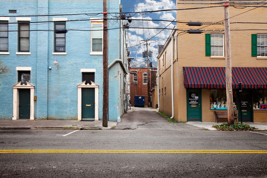 This is a Brian Charles Steel photograph of downtown Cleveland, Tennessee.  A light blue building and a tan building fill the top three quarters of the frame, with an alley and blue sky between them.  The alley has a dark blue dumpster in the back.  A street fills the bottom quarter of the frame with a bright yellow line stretching across the middle of it. 