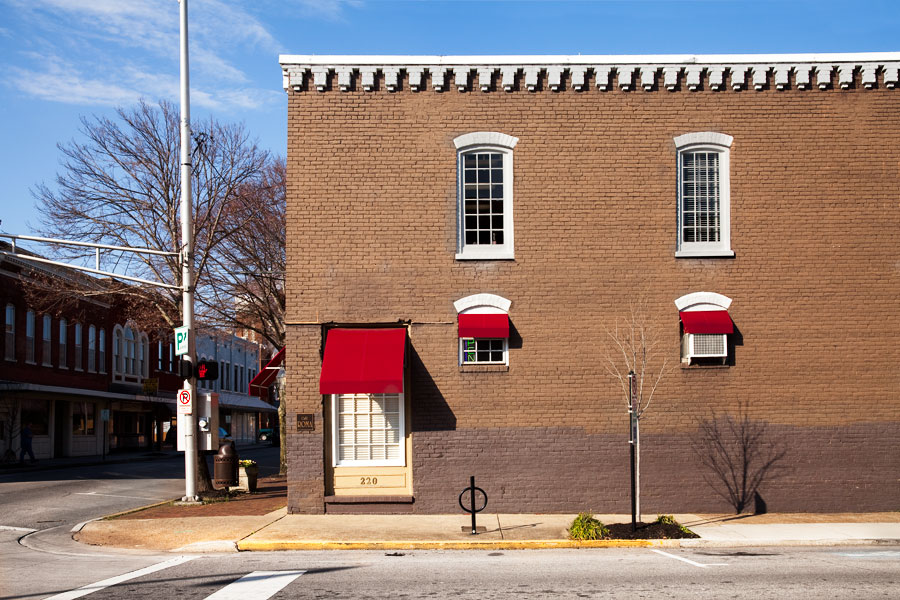 This is a Brian Charles Steel photograph of the outside of Café Roma in Downtown Cleveland, Tennessee.  The building a red brick with white trim.  The windows have white trim and red awnings.  The building takes up the right three fourths of the frame.  The sun beats down on the building while the street to the left of it remains in shadow. 