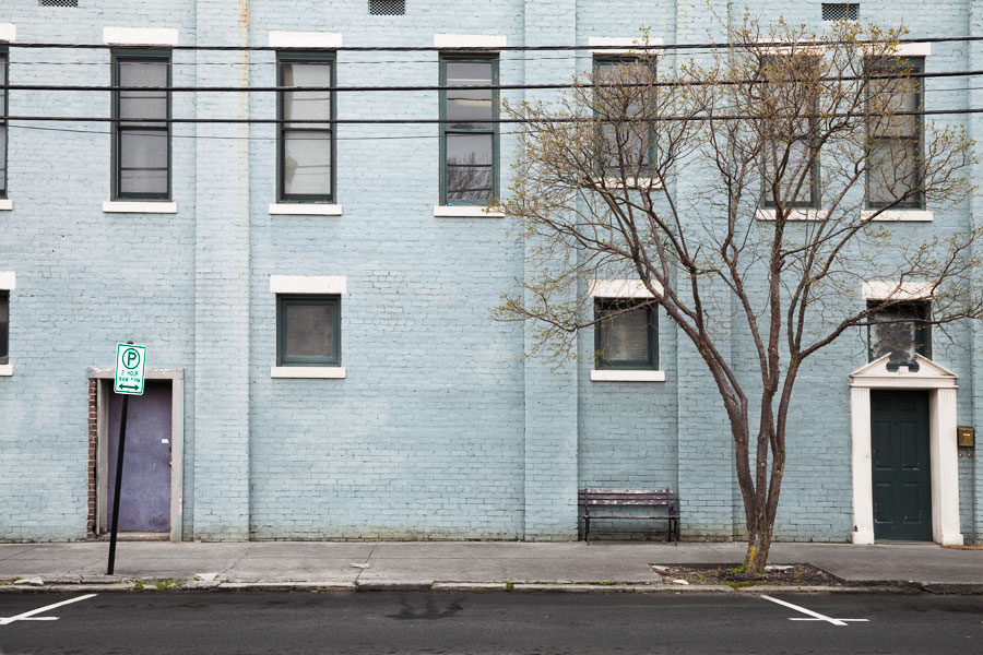 This Brian Charles Steel photograph depicts a light blue building with dark framed windows surrounded by white bricks.  The right half of the frame contains a tree without leaves.  There are protruding rectangles composed of bricks extending from the front of the building.  The rectangles go all the way up the building like pillars.  On the far right side there is a green door surrounded by white trim.  To the left of the door between two brick rectangles is a park bench with worn wood.  On the far left side of the frame is a faded purple door behind a one-hour parking sign. 