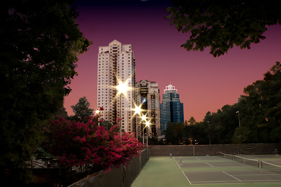 This is a Brian Charles Steel photograph of Park Towers and the King building in Sandy Springs Georgia at sunset.  The towers and king building are in the middle ground.  In the foreground are tennis courts on the right.  On the left in the foreground are trees and bushes with pink flowers.  In the background is an orange and purple sunset sky.  The streetlights in front of the buildings twinkle like stars. 