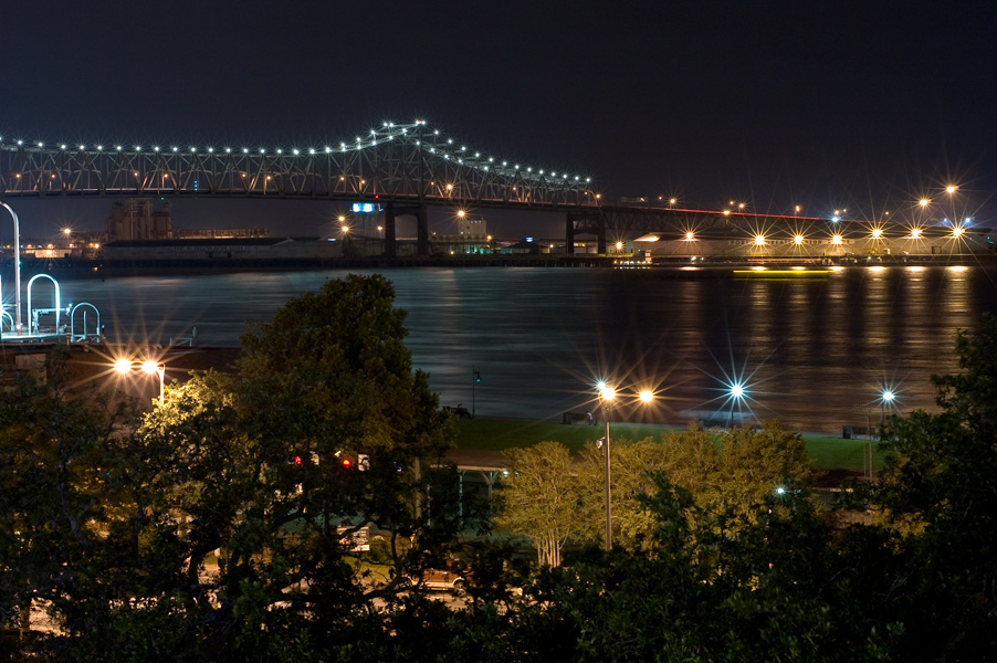 This is a Brian Charles Steel nighttime skyline of the Mississippi river in Baton Rouge Louisiana shot from an elevated position.  The river is in the middle ground.  Lights from the city and the bridge twinkle across the water.  In the foreground are trees and lights.  In the background is city with lights. 