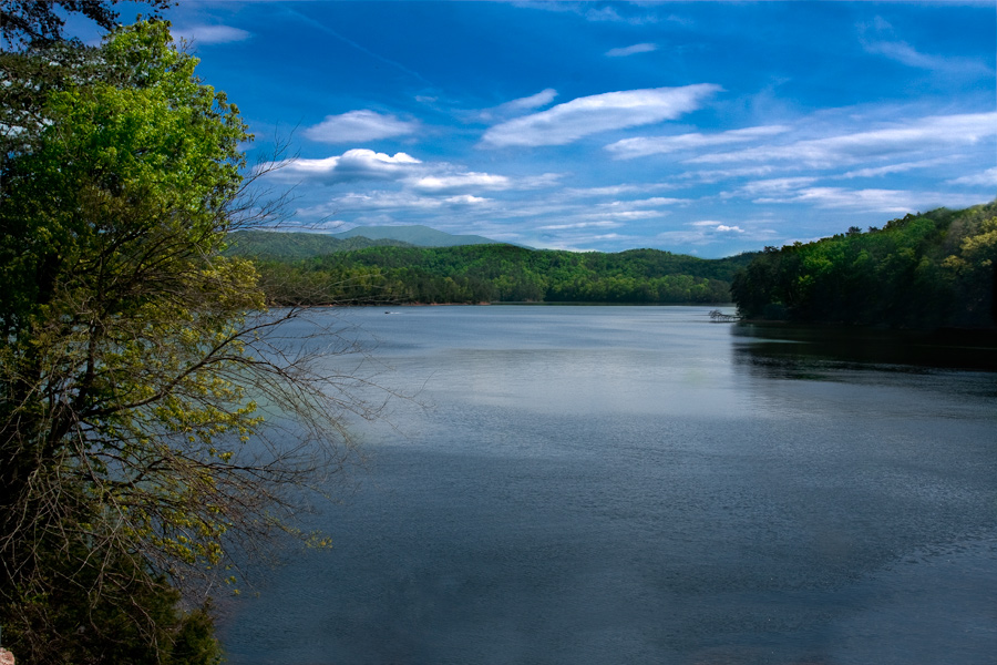 This is a Brian Charles Steel photograph of a lake in Cleveland Tennessee.  The lake composes the lower right portion of the frame.  The upper half of the frame is filled with bright blue sky and white clouds.  Stretching across the middle of the frame are green trees. 