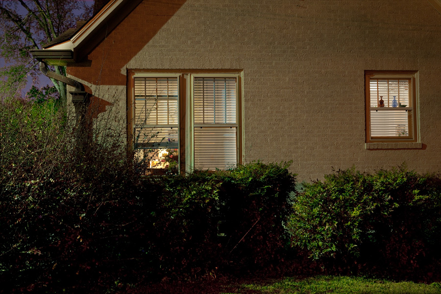 This Brian Charles Steel photograph depicts the outside of a suburban house at night. The outside of the house is tan brick with green gutters and drainpipes. The outside streetlights cast a shadow under the triangular roof.  There are green bushes that go along the house, and come up to the bottom of the window.  The lights are on inside.  In the house you can see a vase of flowers on the dining room table.  