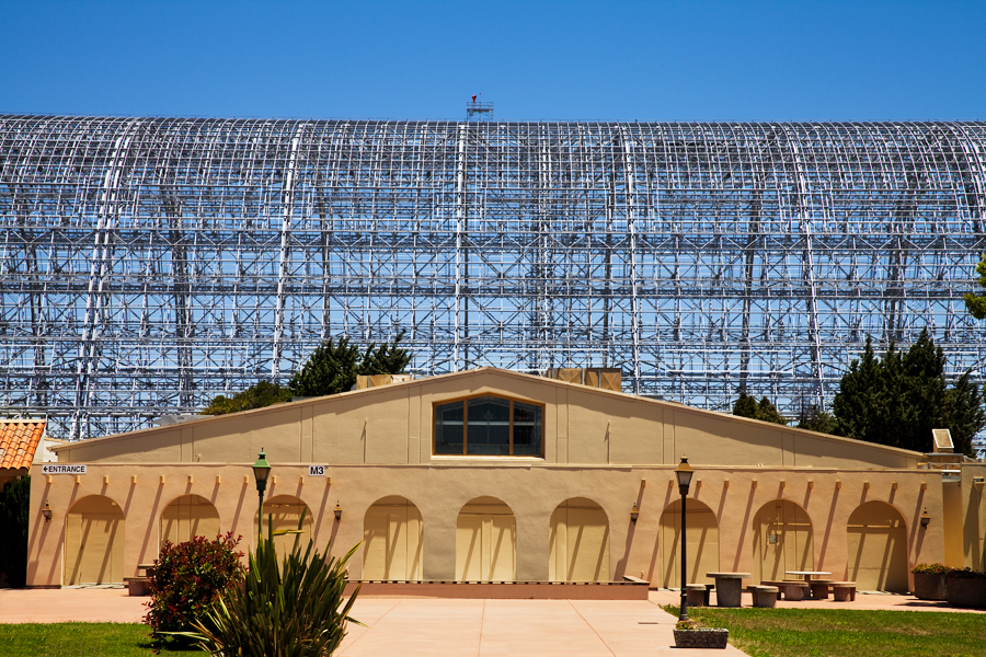 This Brian Charles Steel photo depicts a conference building at NASA Ames Research Center in Mountain View, California.  The building is tan, and has nine arches.  It has a triangular shaped roof with a pentagon shaped window towards the top. The building is in the top of the bottom third of the frame.  Behind the building is a much larger metal frame of a hanger that used to house aircraft.  Behind the hanger is a clear blue sky that takes up about two thirds of the frame.   