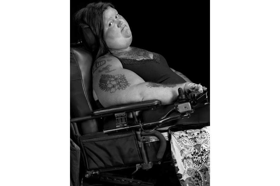 This is a Brian Charles Steel black and white photographic portrait of Jessica Blinkhorn.  She is sitting in her black electric wheelchair.  Her body is facing the right side of the frame at a slight upward angle.  Her head is turned toward the front, and she looks into the camera with a relaxed smile.  She is wearing a dress, and her arms are covered with tattoos. The background is solid black. 