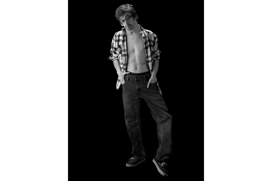 This is a Brian Charles Steel black and white photographic portrait of Barry Lee.  He is standing in the middle of the image and his whole body is in frame.  He is wearing dark jeans and a short sleeve plaid shirt that is unbuttoned, and open.  His hands are in his pockets. He has curly hair and is wearing glasses.  Lee is standing with one leg straight and the other bent.  The background is solid black. 