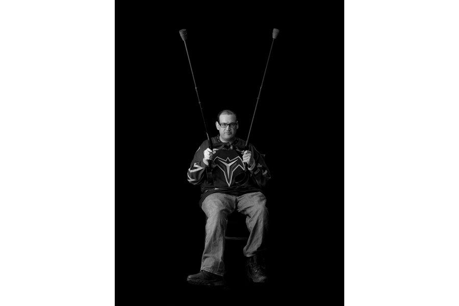 This is a Brian Charles Steel black and white photographic portrait of Ben Kitchings.  Kitchings is in his thirties, and has cerebral palsy.  He is wearing glasses, a Thrashers jersey, and jeans. His whole body is in frame, and he is positioned toward the bottom of the frame.  He holds his crutches up in the air making a v. 