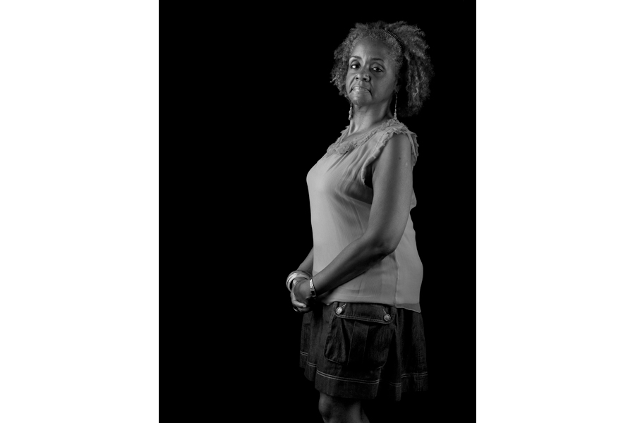 This is a Brian Charles Steel black and white photographic portrait of Candice Holloway.  Candice is positioned in the right half of the frame.  Her hands are folded neatly in front of her, and body is facing to the left while she is looking straight into the camera.  The light is coming from the left. 