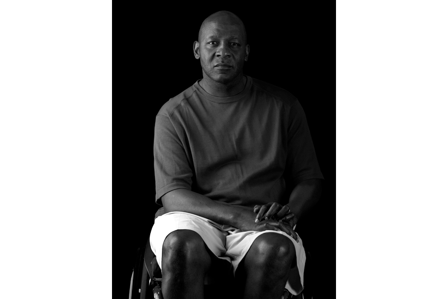 This is a Brian Charles Steel black and white photographic portrait of Curtis Lovejoy.  Lovejoy is wearing a dark short-sleeved shirt and white shorts.  He is seated in his wheelchair and is facing forward.  He is looking straight into the camera and is shown from the knees and up.  His hands are in his lap.  Lovejoy is lit from the left in a Rembrandt style. The background is solid black. 