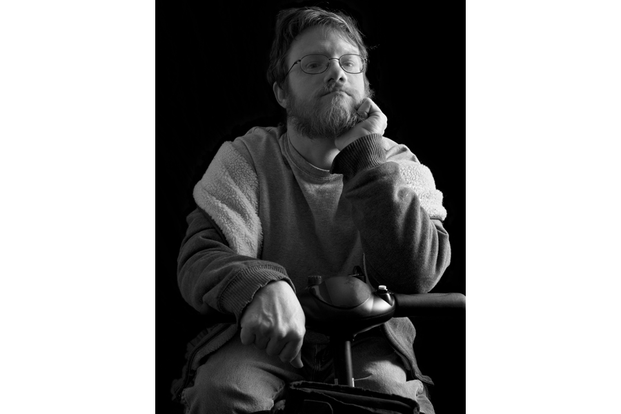 This is a Brian Charles Steel black and white photographic portrait of Eric Carlisle sitting in his scooter.  He is wearing glasses, and looking directly into the camera.  He is wearing a sweater and jeans.  Carlisle is lit in a Rembrandt style with the main light source coming from the right.  He is shown from the knees up, and comprises most of the image. 