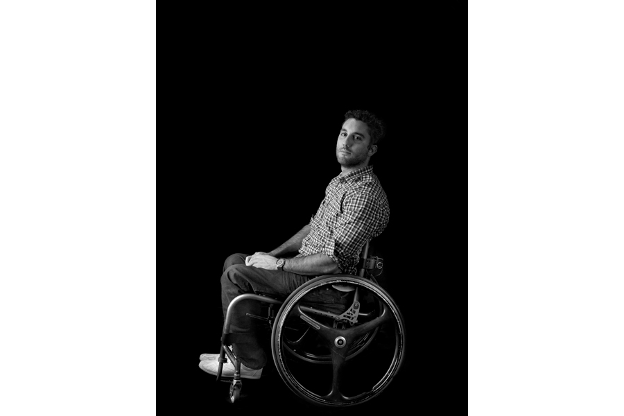 This is a Brian Charles Steel black and white photograph of Kyle Benedict.  He is paraplegic, and sitting in his wheelchair towards the bottom center of the frame.  His chair is facing the left, but he is turned toward the camera looking straight into it.  His hands are folded neatly in his lap.  Benedict has short hair and an athletic build.  The main light source is coming from the left. 
