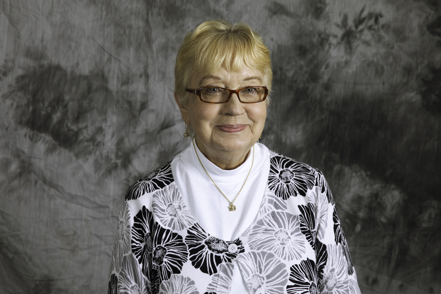 This is a portrait of a woman in front of a grey background. She has short white hair and she is wearing a black and white shirt. 