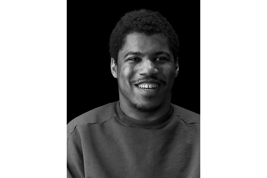 This is a black and white photographic portrait of Charlie taken by Brian Charles Steel.  He is a thirty-year-old African American with cerebral palsy.  He is shown from the chest up, and occupies most of the frame.  It is a close up shot with only his chest and head; his arms are out of frame.  He is looking directly into the camera and smiling.  He is lit in a Rembrandt style with the main light source coming from the left. The background is solid black. 